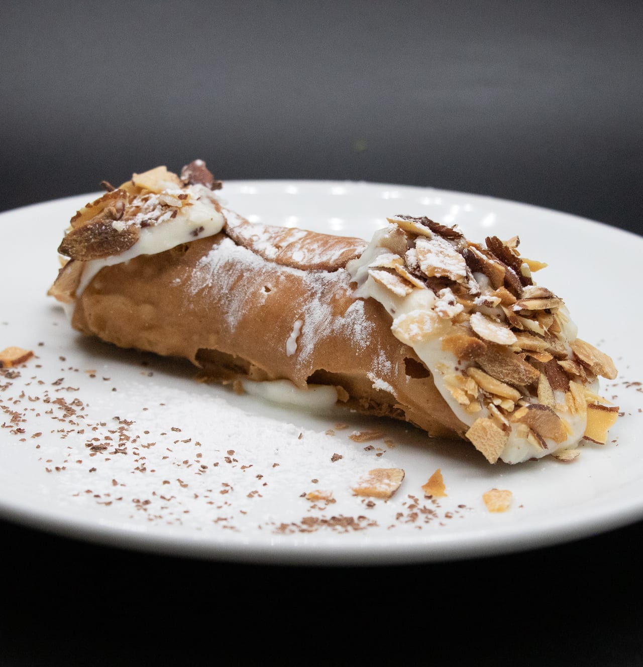 A homemade cannoli on a white plate, dusted with powdered sugar and topped with toasted almonds
